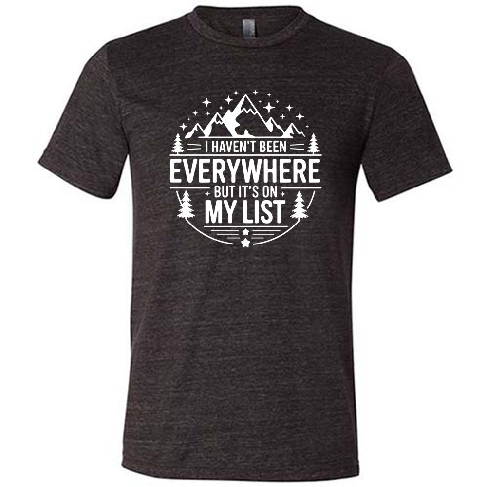 black unisex tee with the words "I haven't been everywhere but it's on my list" as part of a circular design involving the graphic of small mountains on top and a few trees on the sides and stars scattered around