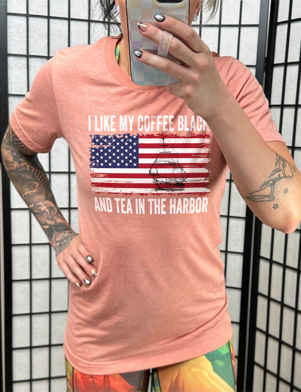 sunset unisex tee with the words "I like my coffee black and tea in the harbor" on it, with a graphic in the center of an American Flag, and the sketch of a masted ship on it