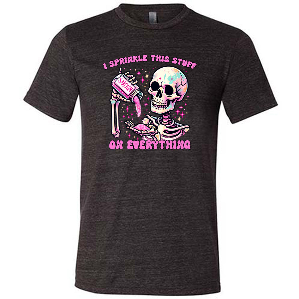 black unisex tee with the words "I sprinkle this stuff on everything" on it, and the graphic of a skeleton who is pouring a jar labeled "sarcasm" filled with pink glittery material into its hand, surrounded by little pink and white stars