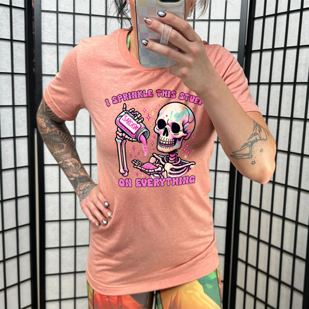 sunset unisex tee with the words "I sprinkle this stuff on everything" on it, and the graphic of a skeleton who is pouring a jar labeled "sarcasm" filled with pink glittery material into its hand, surrounded by little pink and white stars
