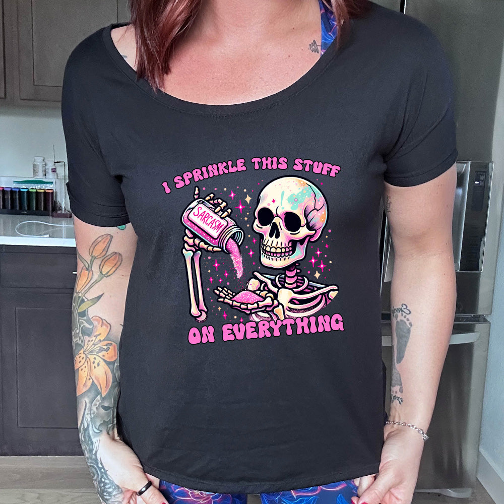black slouchy tee with the words "I sprinkle this stuff on everything" on it, and the graphic of a skeleton who is pouring a jar labeled "sarcasm" filled with pink glittery material into its hand, surrounded by little pink and white stars