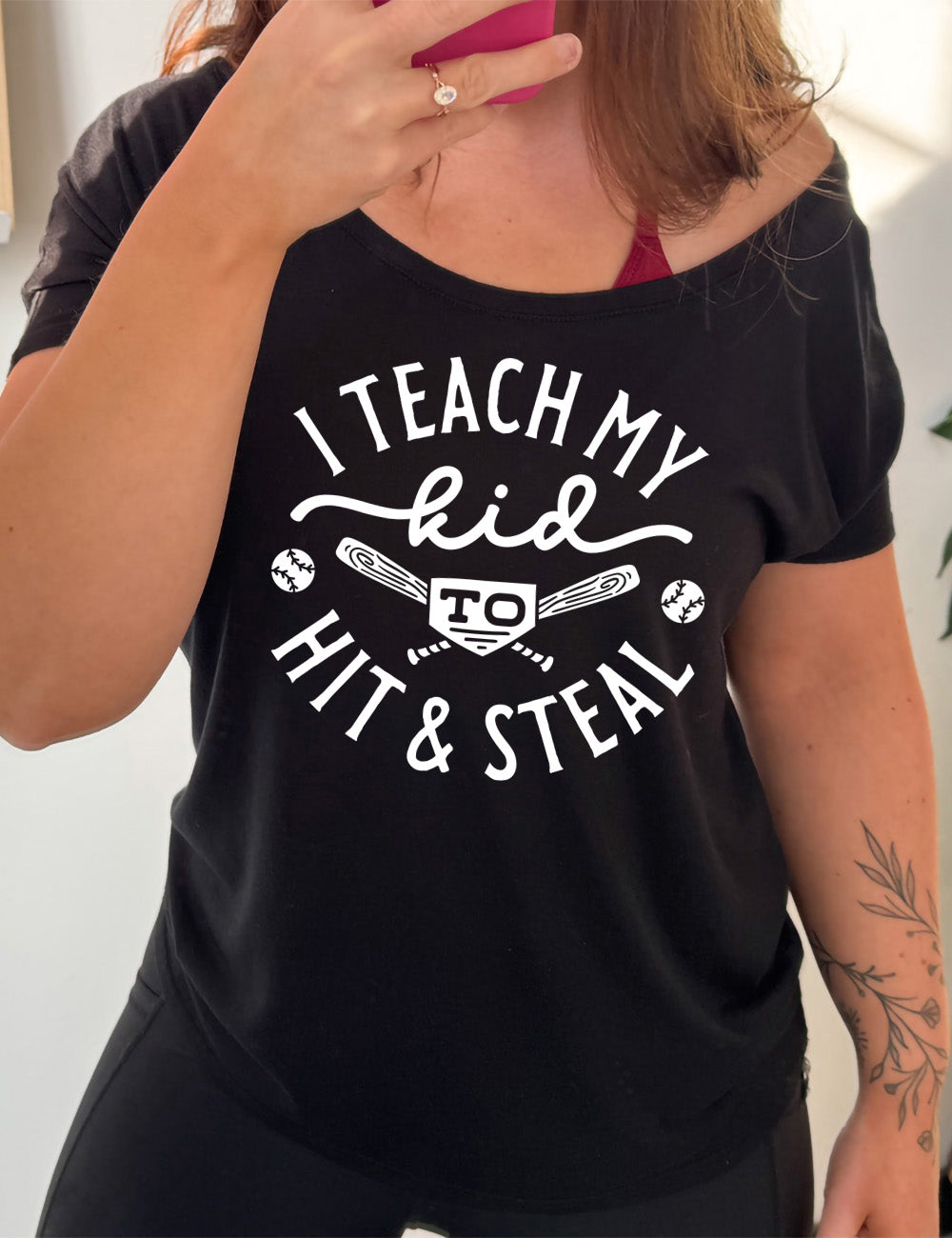 black slouchy tee with words saying "I teach my kid to hit & steal" on it, with two bats crossed behind the word "to", and a baseball on either side of that
