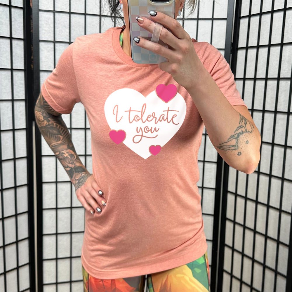 sunset unisex tee with the words "I tolerate you" in sunset lettering on it, within a white heart graphic that has three small pink hearts overlapping on its edges 
