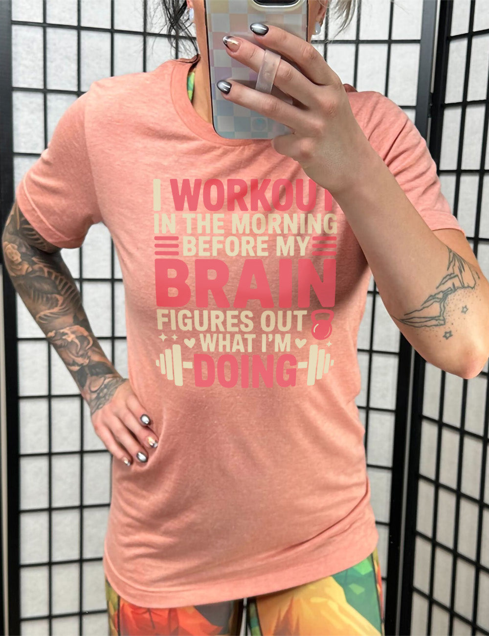 sunset unisex tee with words on it saying "I workout in the morning before my brain figures out what I'm doing", with the graphics of lines, a kettlebell, a couple stars and hearts, and a barbell
