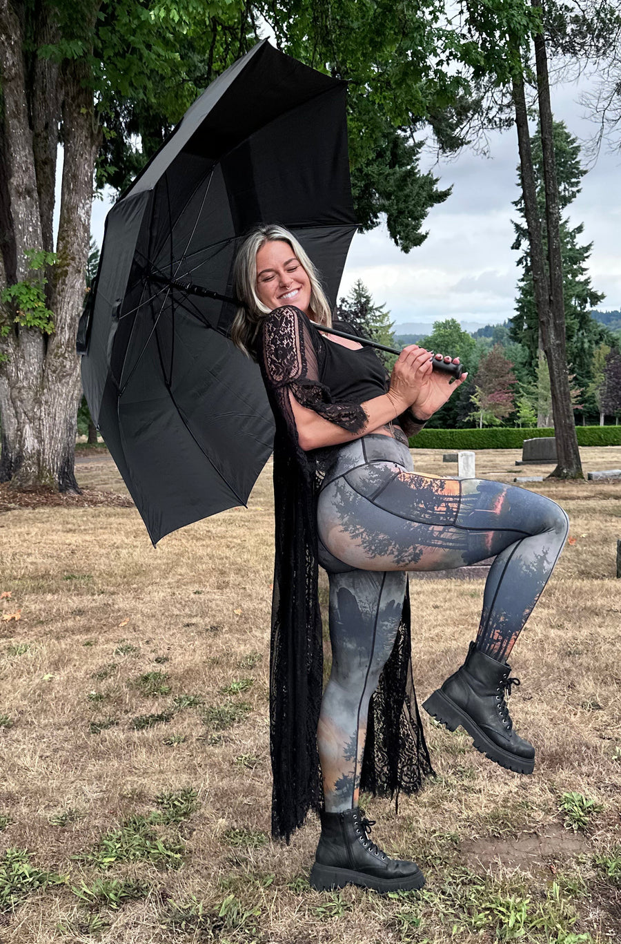A woman posing in a cemetery holding a large black umbrella behind her. She is wearing a black top, distressed blue jeans with a graphic print, and black combat boots.