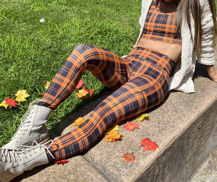 A person sitting outdoors on a stone ledge wearing a matching orange, black, and black plaid crop top and leggings set, with white lace-up ankle boots and a white textured cardigan, with scattered autumn leaves on the grass and stone.