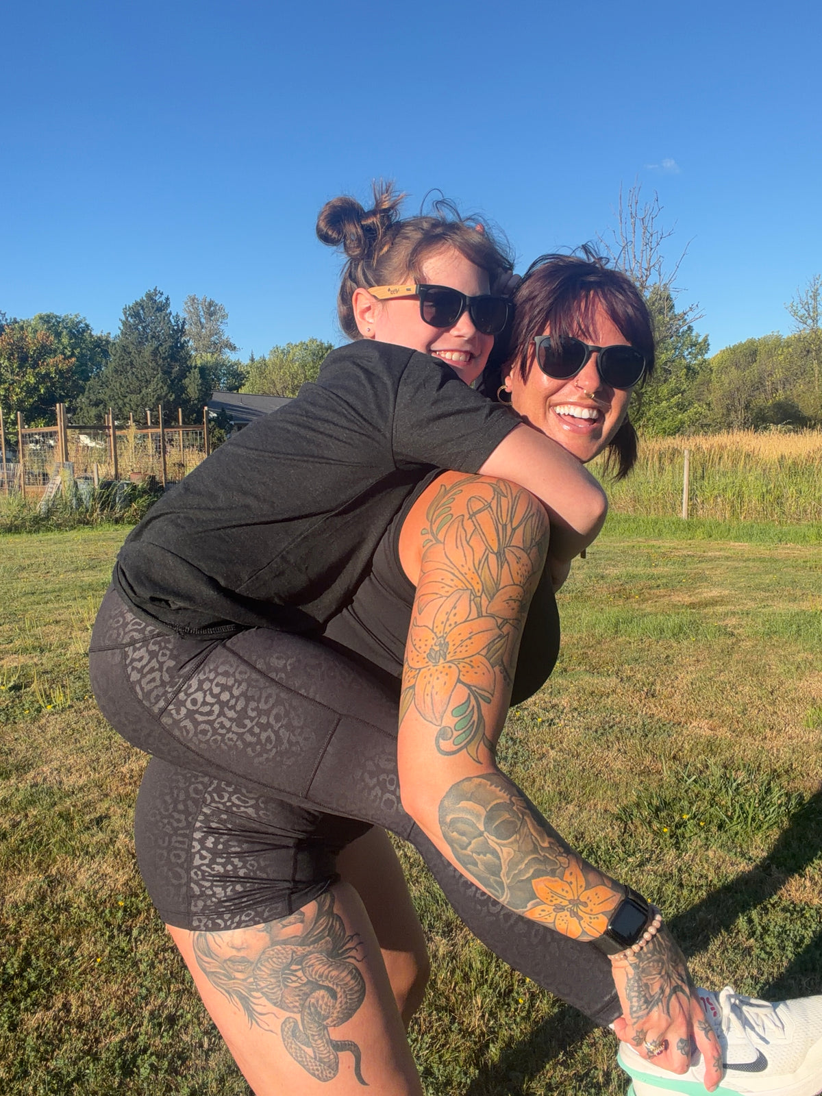 Two women outdoors in a grassy field, with one piggybacking on the other. The woman being carried wears a black t-shirt, black leopard leggings, and white sneakers, while the carrier wears a black tank top and black leopard shorts.