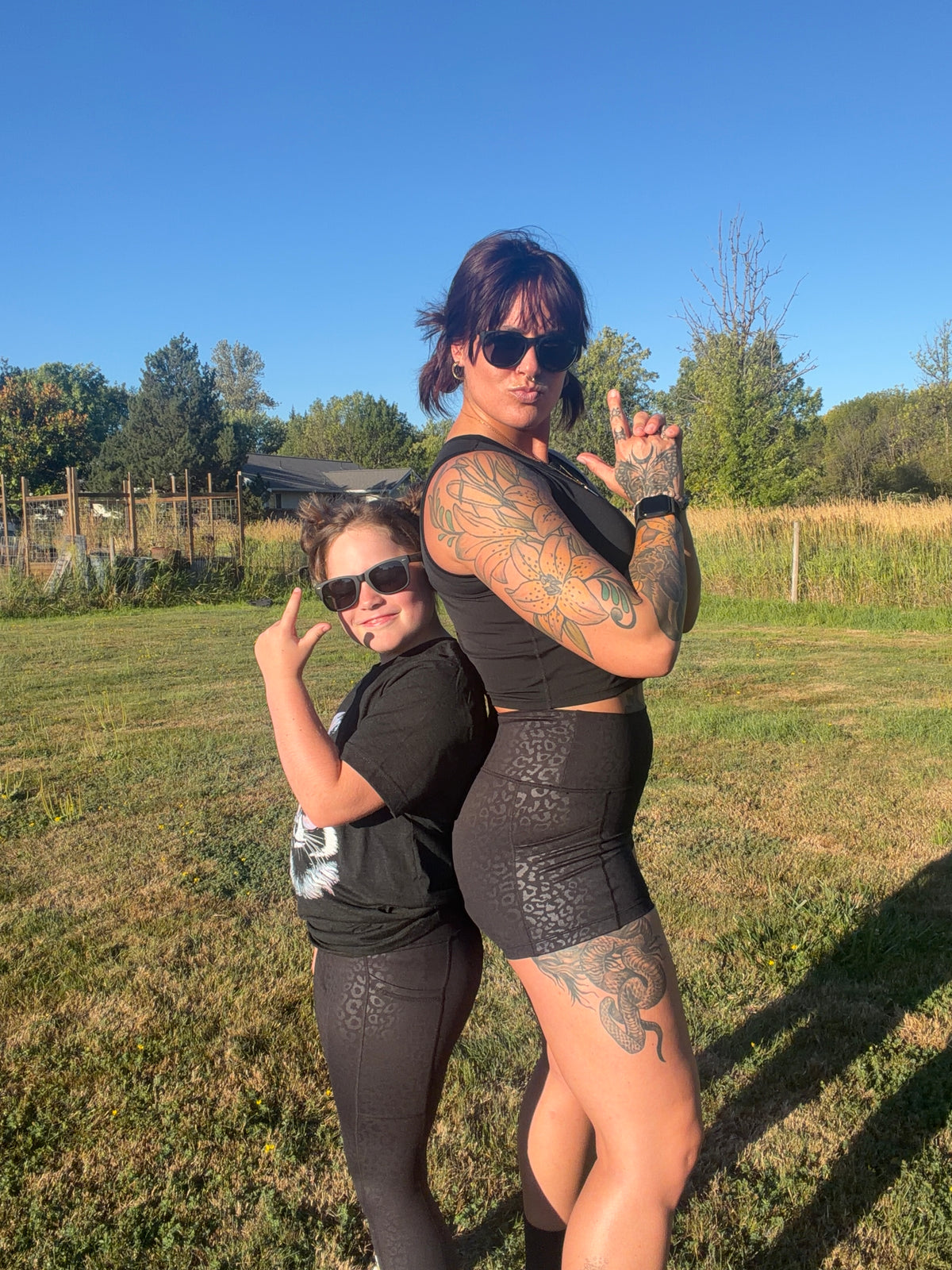 Two women posing back-to-back outdoors on a grassy field under a clear blue sky, wearing matching black activewear sets.