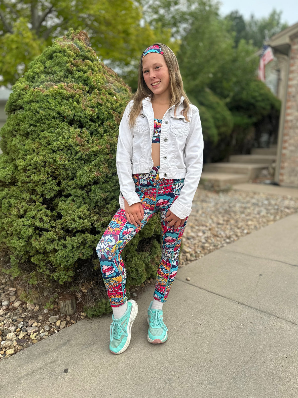 A young woman stands outdoors on a concrete walkway wearing a matching colorful graphic print activewear set consisting of a crop top and leggings, a white cropped denim jacket, and mint green sneakers.