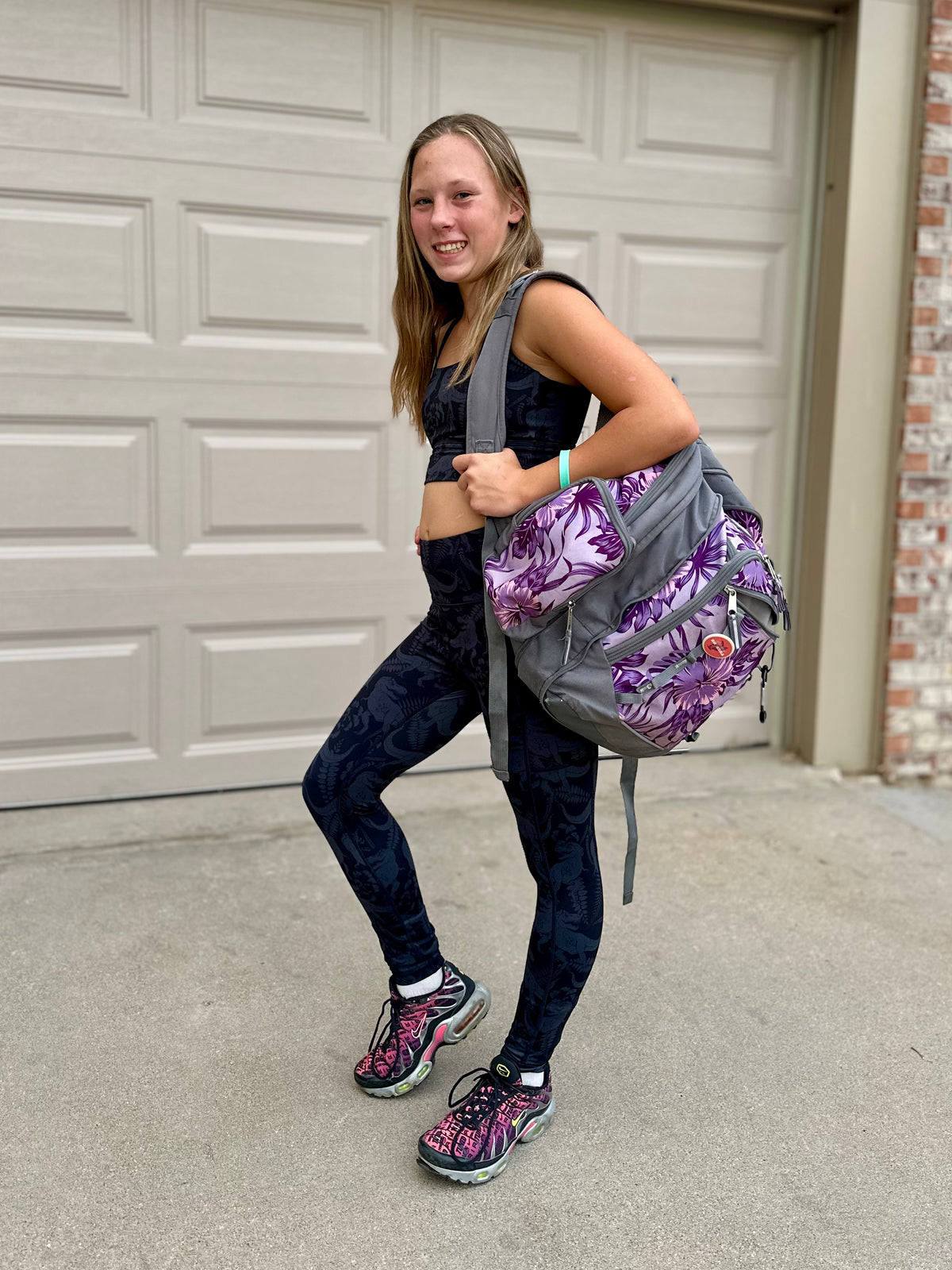 A woman wearing a black sports bra and dark patterned leggings carries a gray backpack with a purple floral print, standing in front of a white garage door.