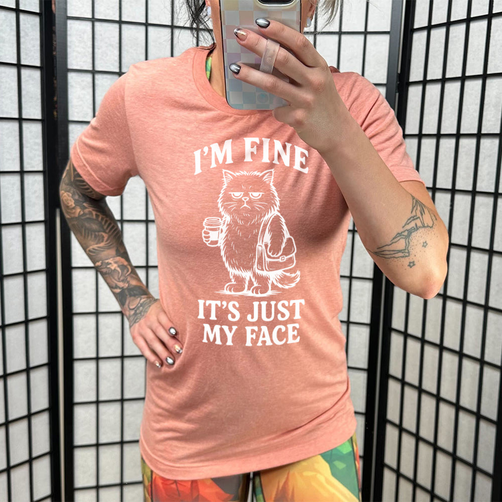 sunset unisex tee with the words "I'm fine it's just my face" on it, around the graphic of a cat with a resting-grumpy face, holding a coffee and wearing a purse