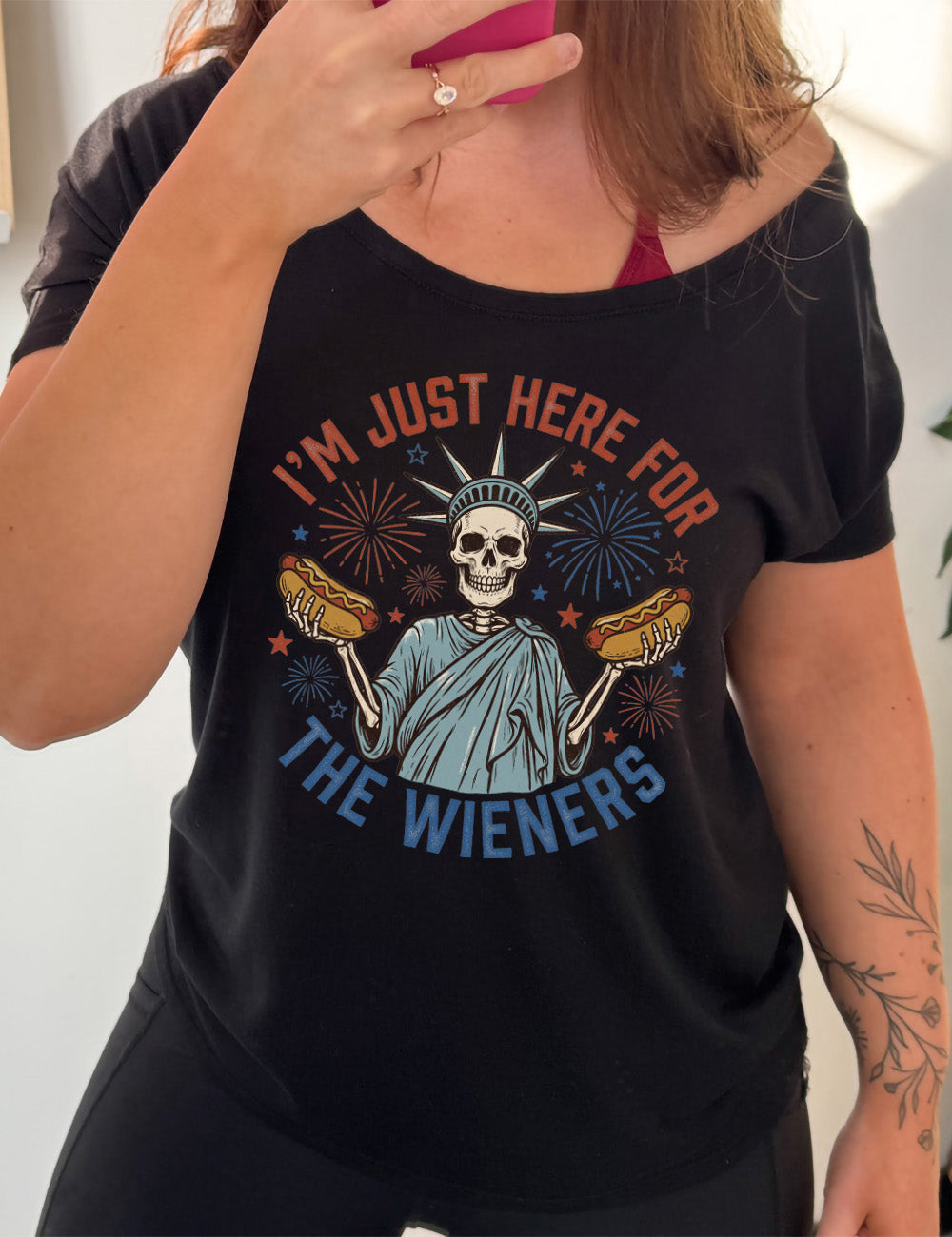 Black slouchy tee with the words "I'm just here for the wieners" on it, with the graphic in the middle of the words of a skeletal statue of liberty with a hotdog in each hand and fireworks and stars around it