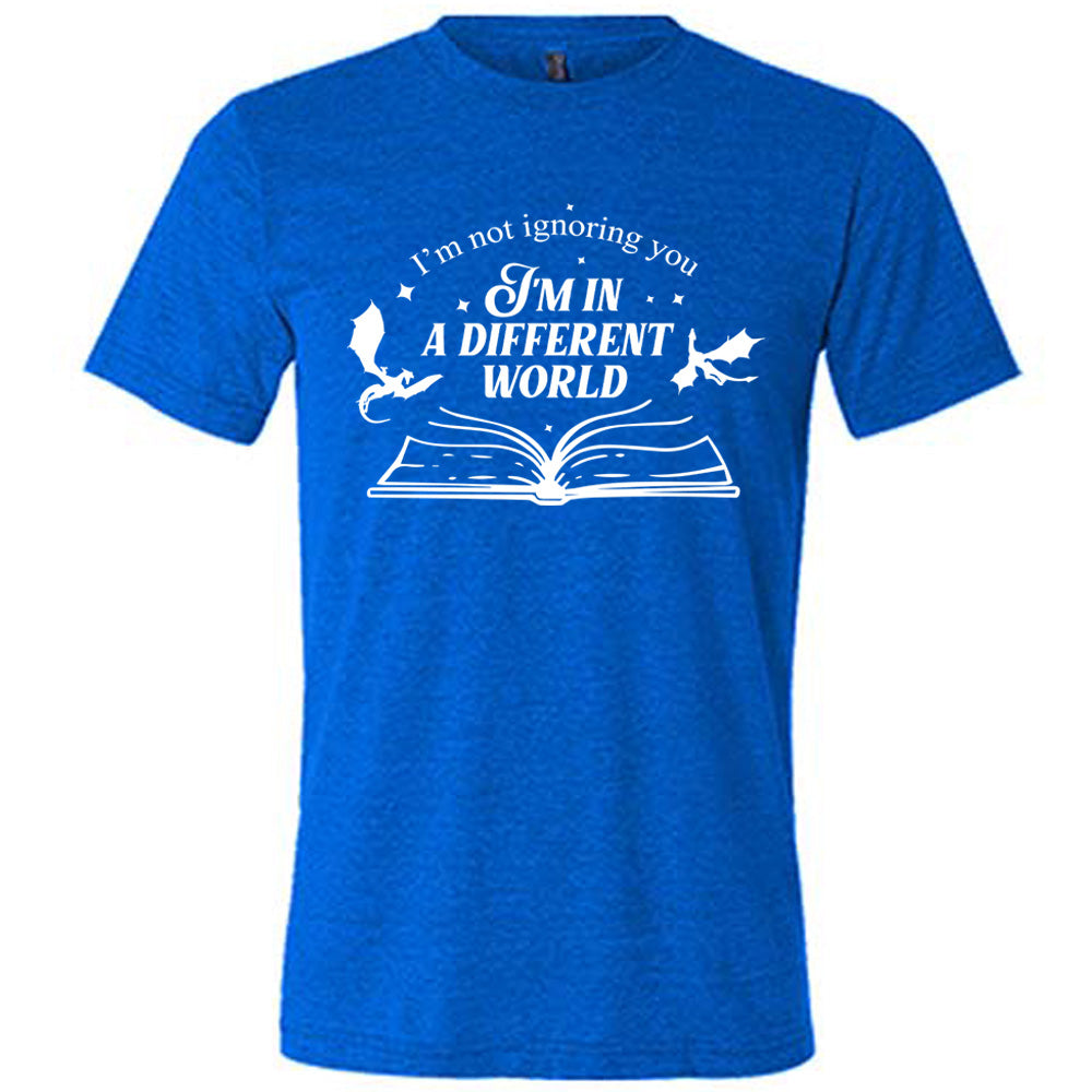blue unisex tee with the words "I'm not ignoring you I'm in a different world" on it, with the graphic of a large book, lying open, and two little dragons flying next to the words, a few stars scattered around