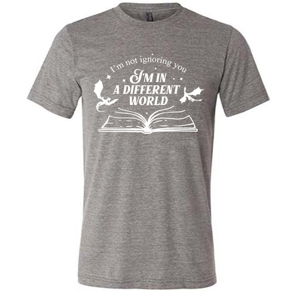 grey unisex tee with the words "I'm not ignoring you I'm in a different world" on it, with the graphic of a large book, lying open, and two little dragons flying next to the words, a few stars scattered around