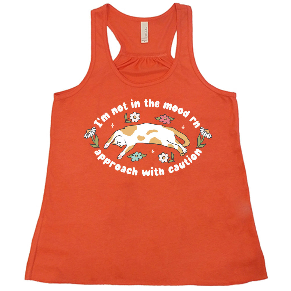 coral racerback tank with the words "I'm not in the mood rn approach with caution" on it in an oval, with the graphic of a cat splayed on its back within the words, and a few flowers around