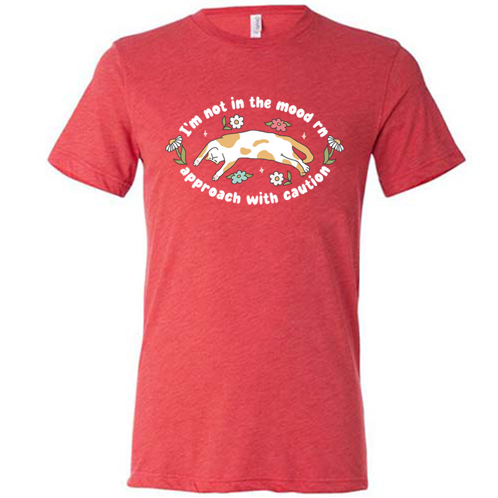 red unisex tee with the words "I'm not in the mood rn approach with caution" on it in an oval, with the graphic of a cat splayed on its back within the words, and a few flowers around