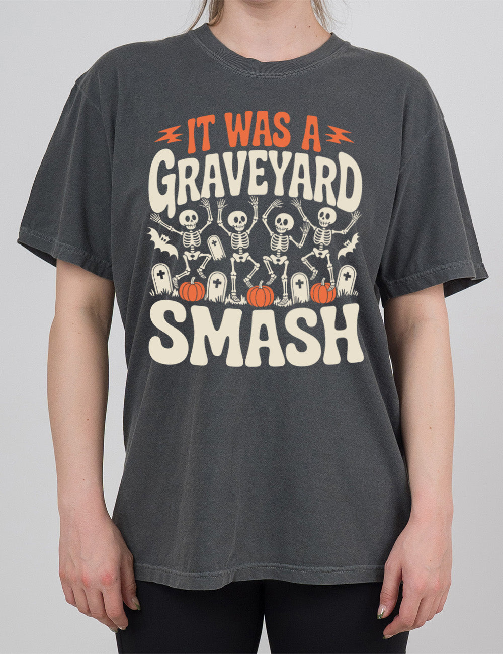 pepper modern tee with the words "it was a graveyard smash" with a lightning bolt on either side of the words "it was a" and the large graphic in the middle of the words of four skeletons dancing next to tombstones, pumpkins, and two bats