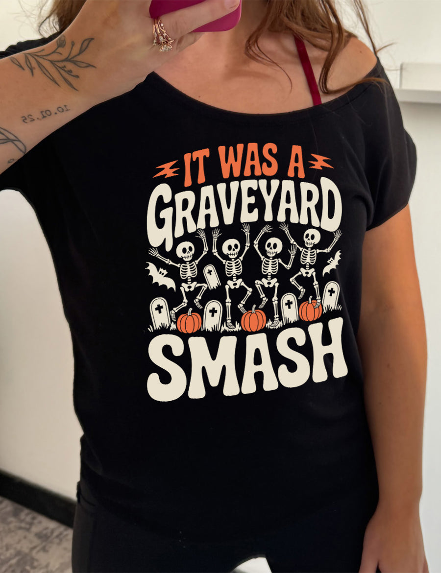 black slouchy tee with the words "it was a graveyard smash" with a lightning bolt on either side of the words "it was a" and the large graphic in the middle of the words of four skeletons dancing next to tombstones, pumpkins, and two bats