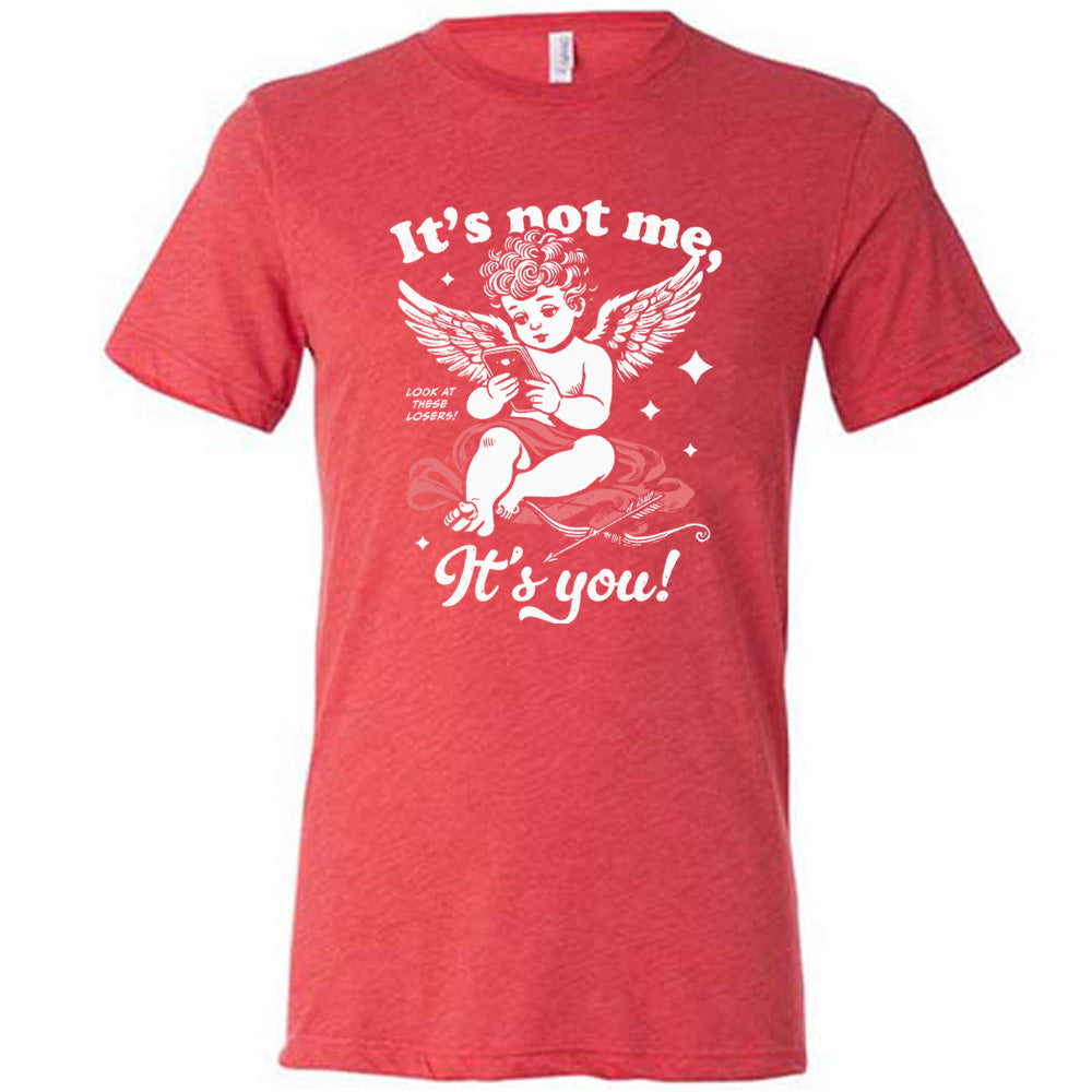 red unisex tee with the words "it's not me, it's you!" on it, with graphics of Cupid looking at a cell phone (presumably looking at dating apps), with small words saying "look at these losers!" and Cupid's bow and arrow at his side