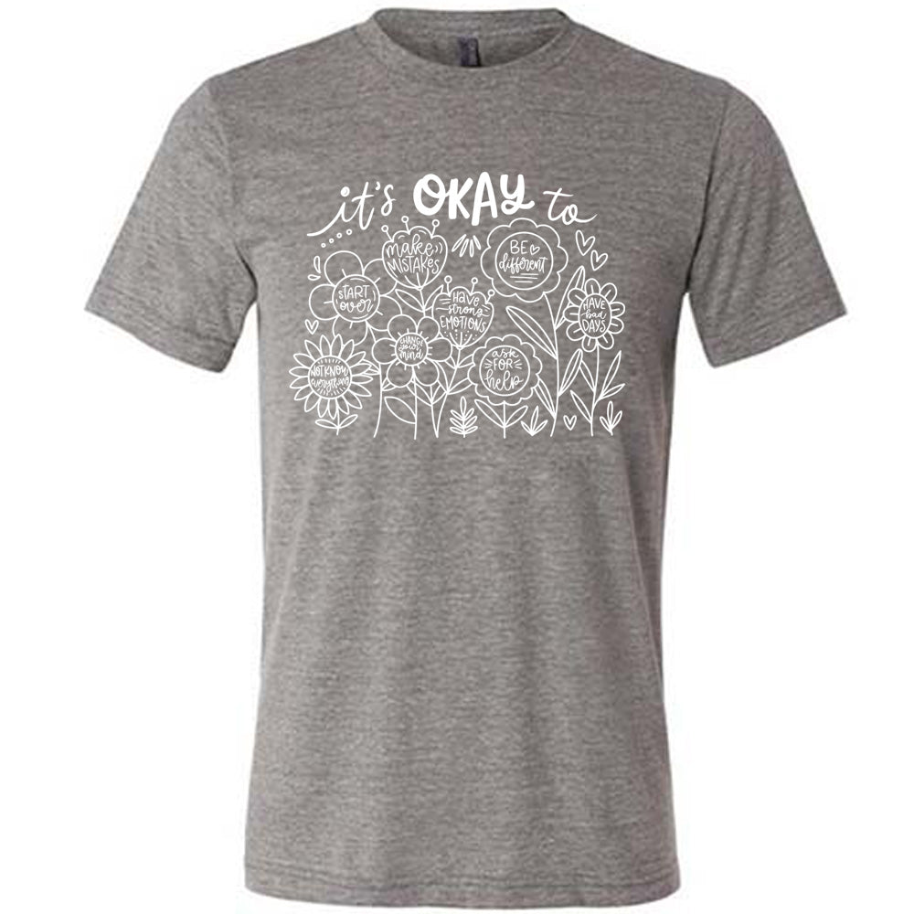 grey unisex tee with the words "it's okay to" on it, and underneath there are many flowers that each have the completion of that phrase; the different endings are "make mistakes", "be different", "have bad days", "ask for help", "have strong emotions", "change your mind", "Start over", and "not know everything".