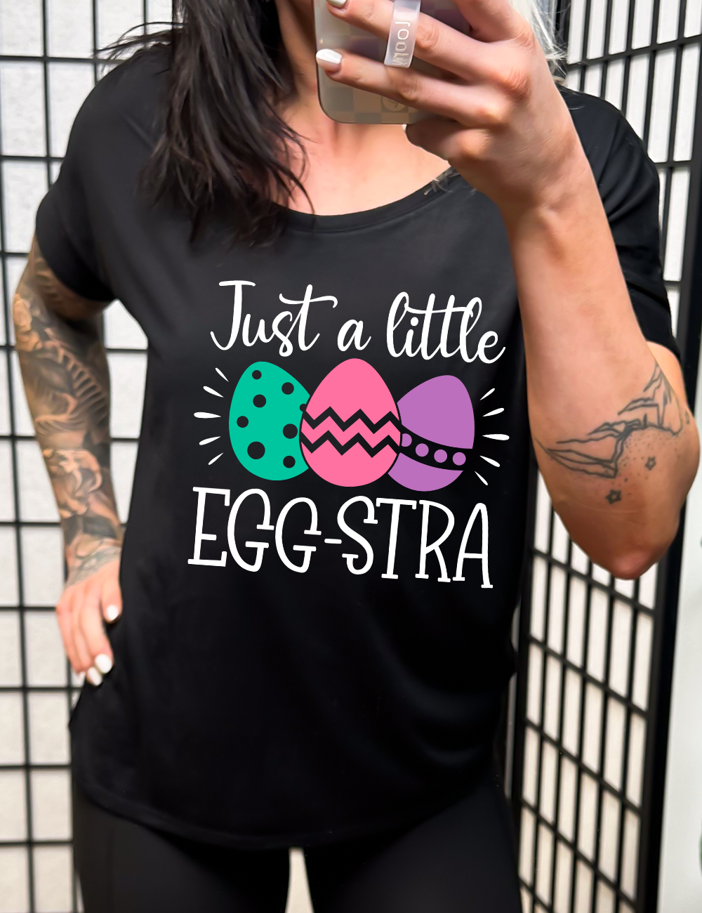 black slouchy tee with the words "just a little egg-stra" on it, with the graphic of three decorated easter eggs 