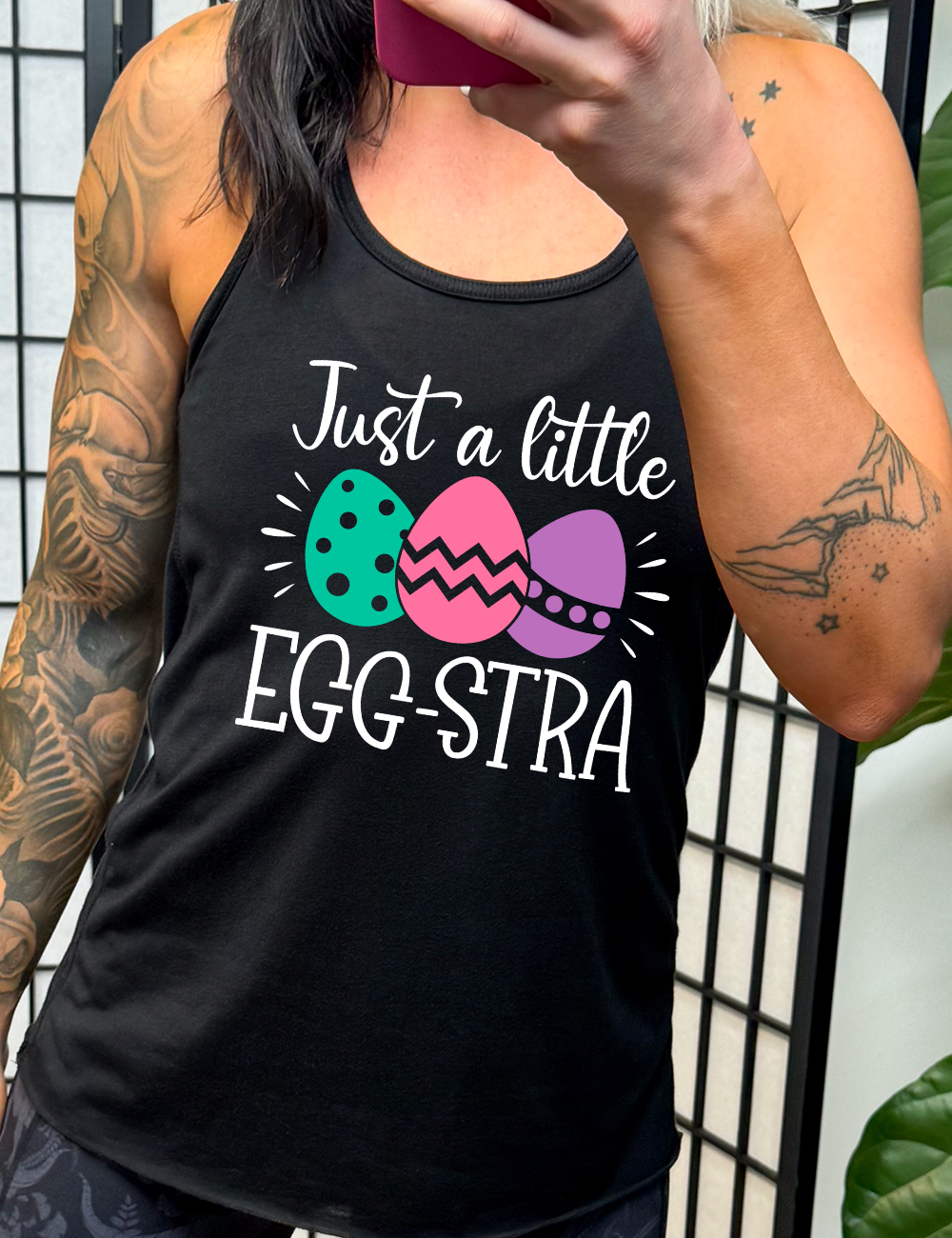 black racerback tank with the words "just a little egg-stra" on it, with the graphic of three decorated easter eggs