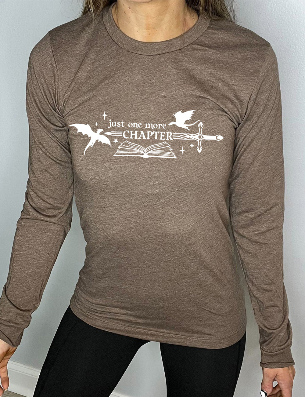 heather brown long sleeve shirt with the graphic of an open book, with stars and a couple dragon silhouettes, and a sword, and the words "just one more chapter" on it, with the word "chapter" overlaying the sword