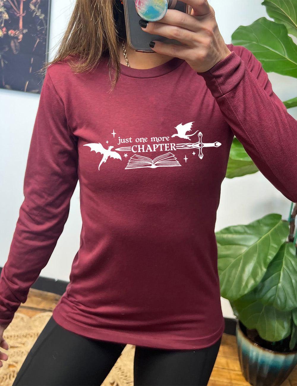 heather cardinal long sleeve shirt with the graphic of an open book, with stars and a couple dragon silhouettes, and a sword, and the words "just one more chapter" on it, with the word "chapter" overlaying the sword
