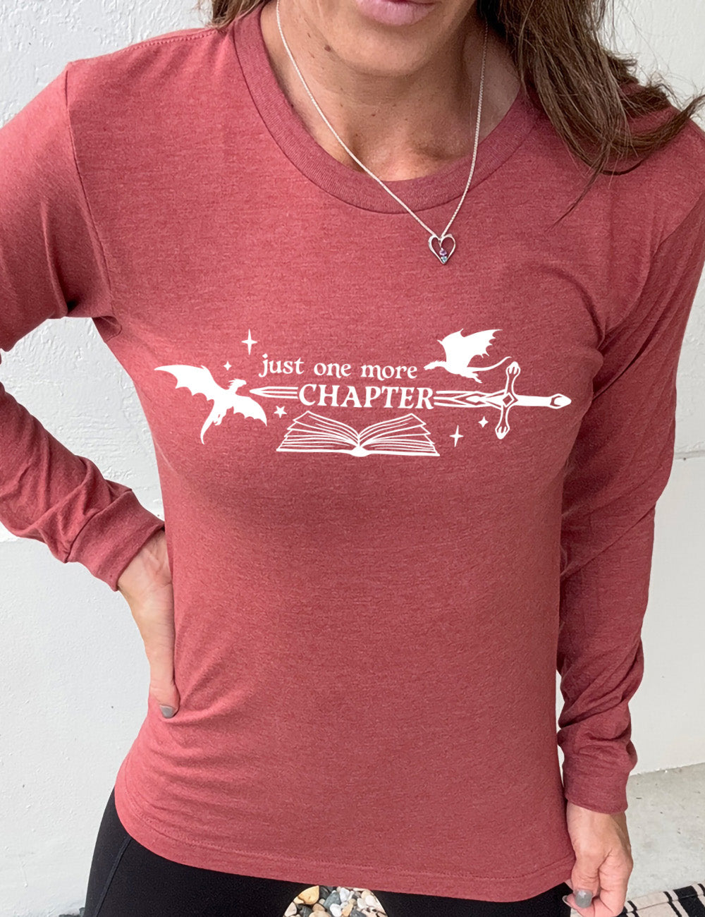 heather clay long sleeve shirt with the graphic of an open book, with stars and a couple dragon silhouettes, and a sword, and the words "just one more chapter" on it, with the word "chapter" overlaying the sword