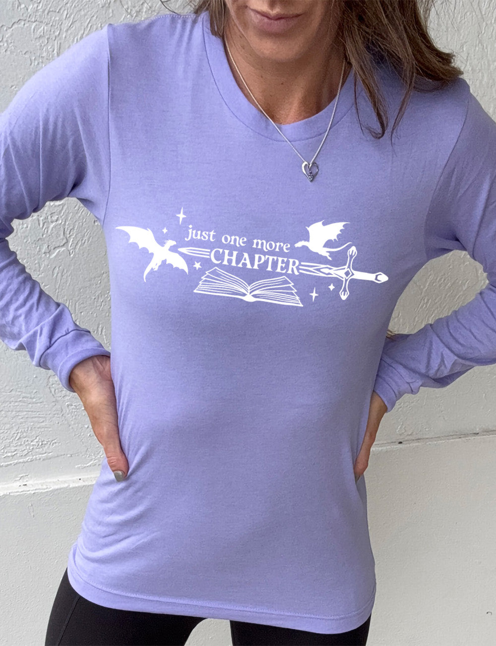 heather dark lavender long sleeve shirt with the graphic of an open book, with stars and a couple dragon silhouettes, and a sword, and the words "just one more chapter" on it, with the word "chapter" overlaying the sword