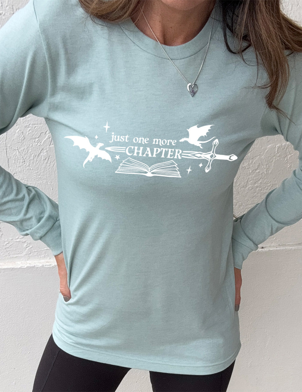 heather dusty blue long sleeve shirt with the graphic of an open book, with stars and a couple dragon silhouettes, and a sword, and the words "just one more chapter" on it, with the word "chapter" overlaying the sword