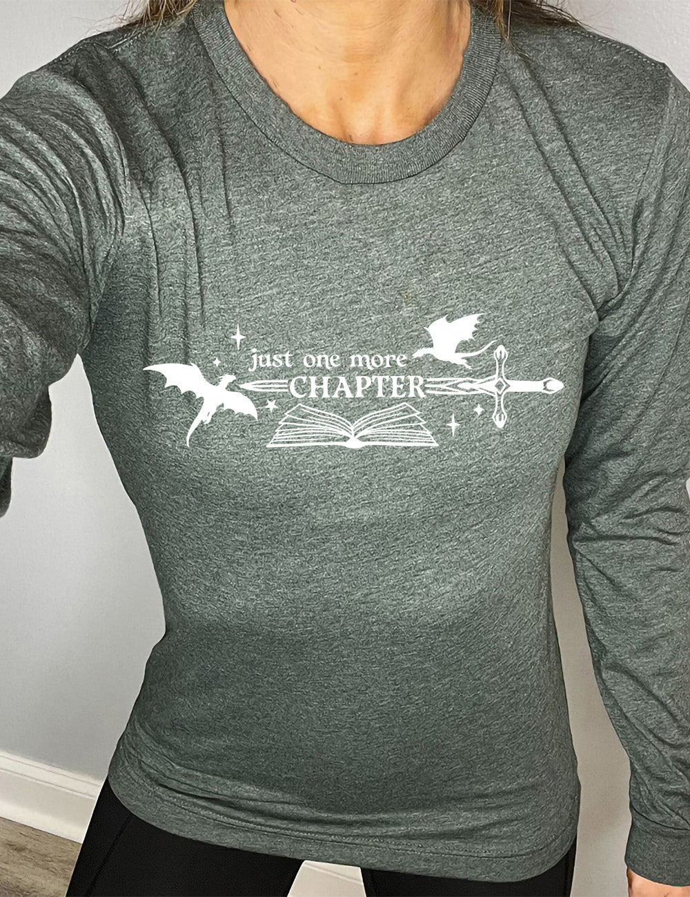 heather forest long sleeve shirt with the graphic of an open book, with stars and a couple dragon silhouettes, and a sword, and the words "just one more chapter" on it, with the word "chapter" overlaying the sword
