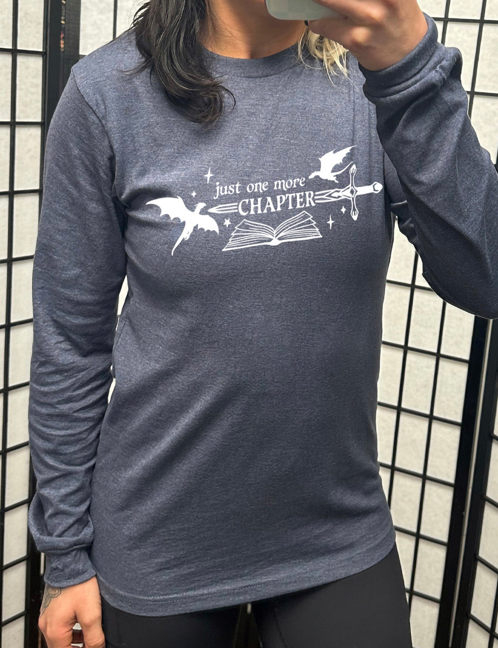 heather navy long sleeve shirt with the graphic of an open book, with stars and a couple dragon silhouettes, and a sword, and the words "just one more chapter" on it, with the word "chapter" overlaying the sword