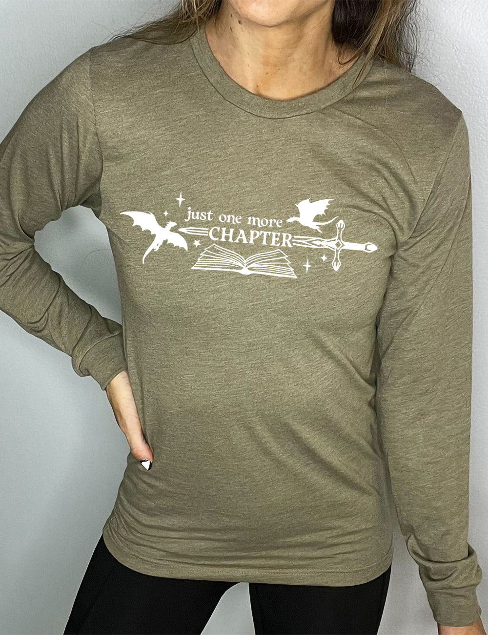 heather olive long sleeve shirt with the graphic of an open book, with stars and a couple dragon silhouettes, and a sword, and the words "just one more chapter" on it, with the word "chapter" overlaying the sword