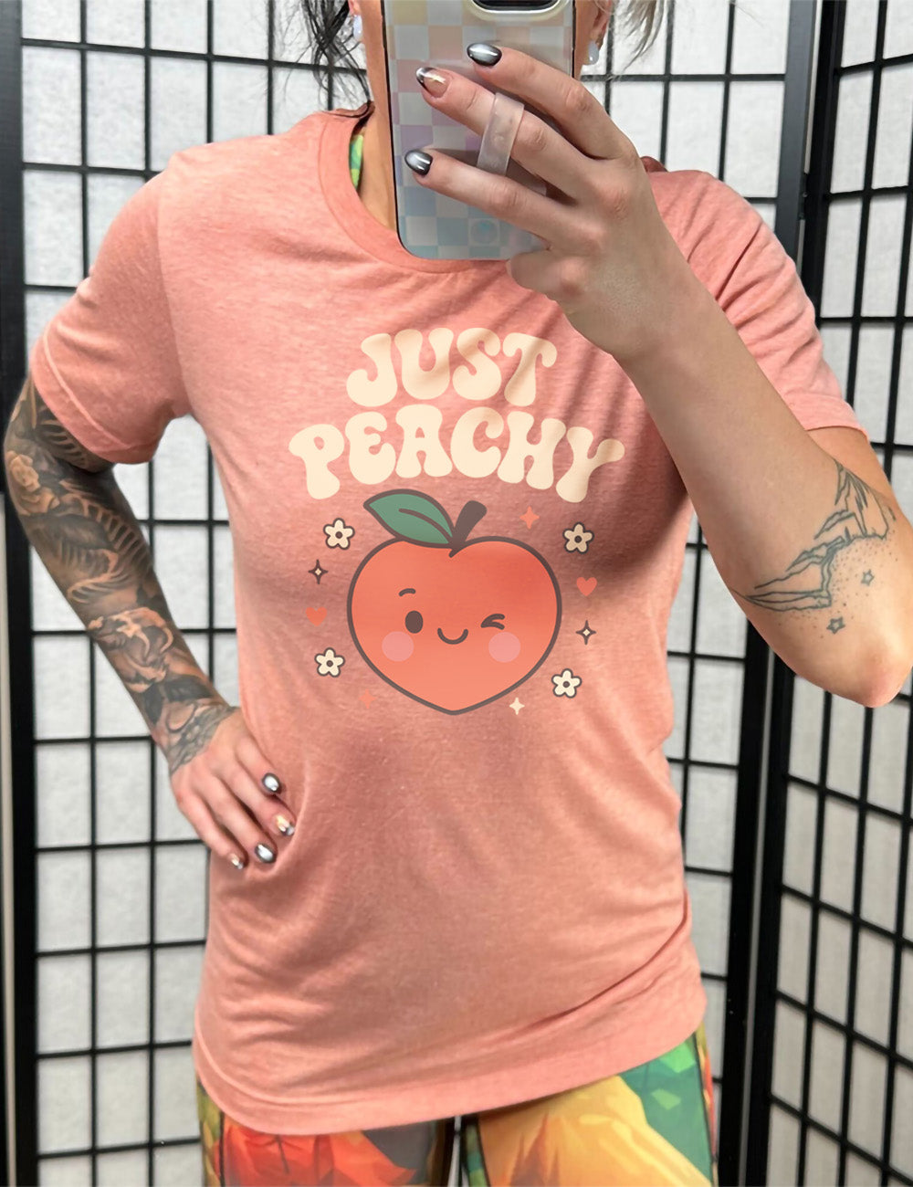sunset unisex tee with the graphic of a winking peach, surrounded by little hearts, flowers, and stars, with the words above it saying "just peachy"