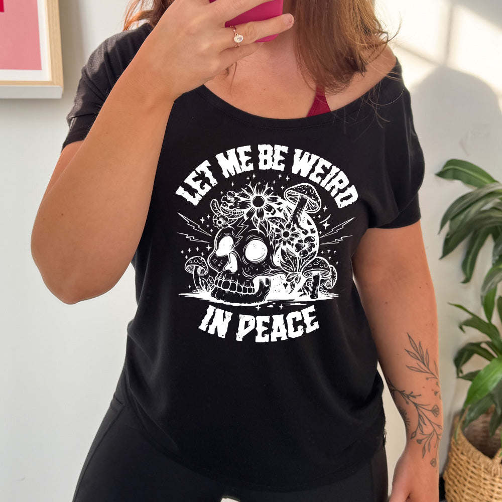 black slouchy tee with the words "let me be weird in peace" on it with the graphic of a skull on the ground with mushrooms growing on and around it, as well as flowers growing on it, surrounded by little stars and lightening bolts