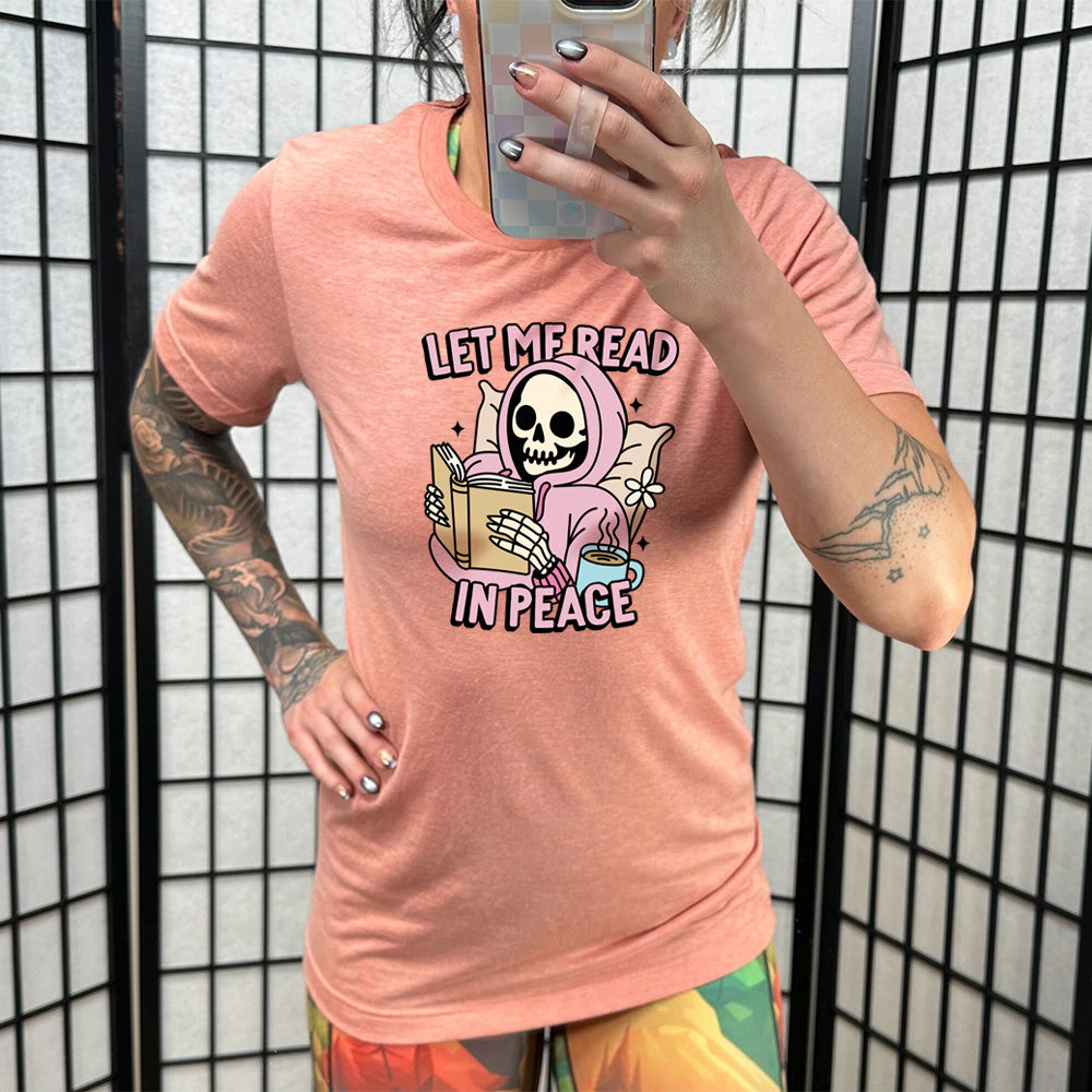 coral shirt with the text "Let Me Read In Peace" and a skeleton reading graphic on it