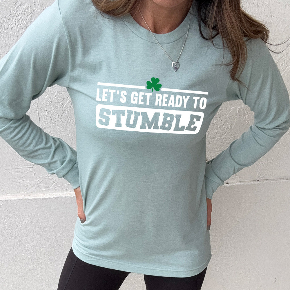 heather dusty blue long sleeve with the words "let's get ready to stumble" on it, with one little shamrock graphic, and the word "stumble" in a rectangular block, and the letters leaning in different directions