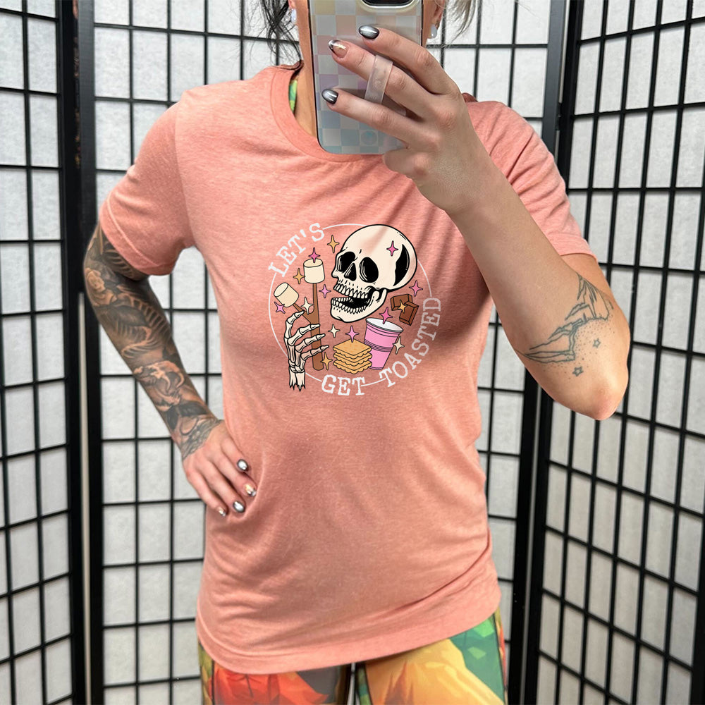 Sunset colored shirt with a skull and marshmallow stick graphic on it that says "Let's Get Toasted" 