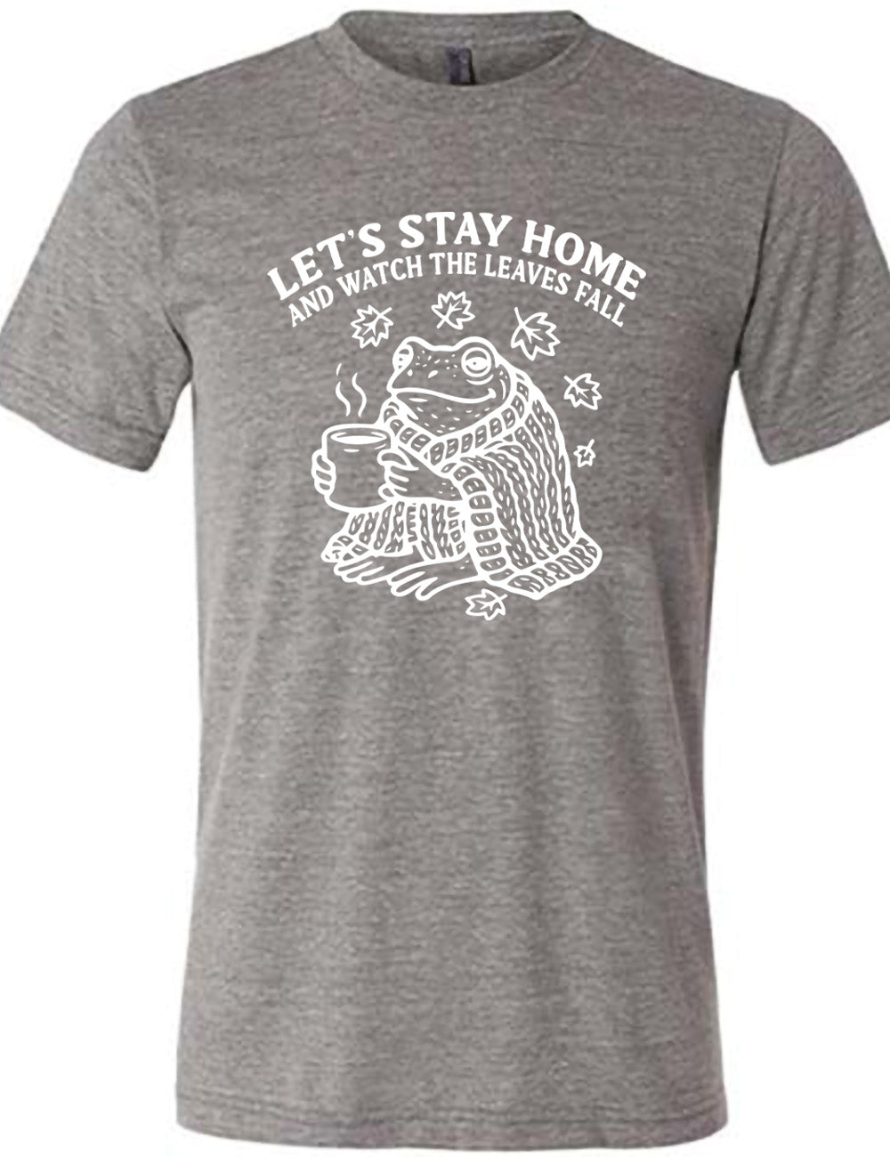 grey unisex tee with the words "let's stay home and watch the  leaves fall" on it, and the graphic underneath of a frog, wrapped in a blanket, holding a mug of steaming beverage, with five leaves arching over it