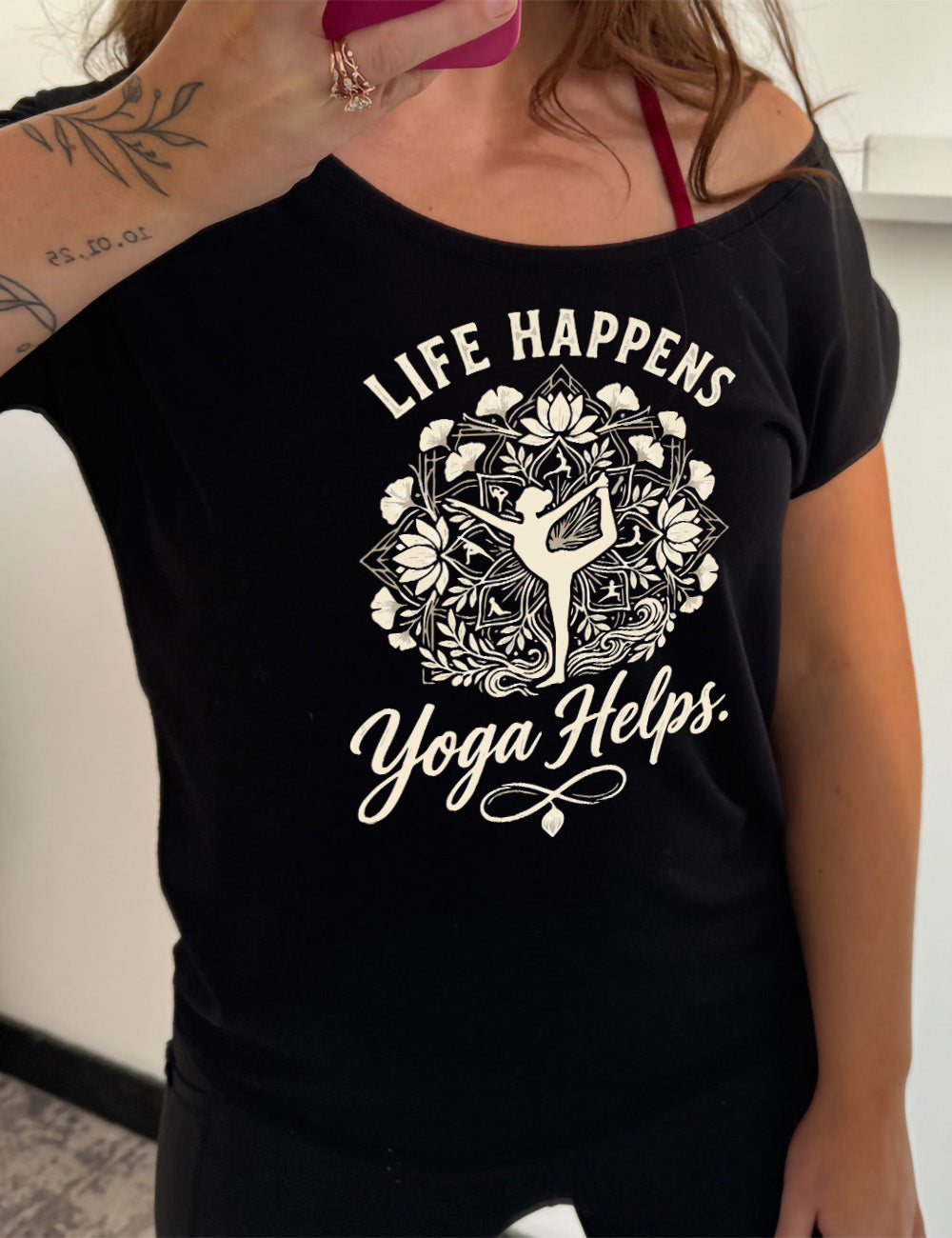 black slouchy tee with the words "life happens yoga helps." on it, around the graphic of a woman's silhouette doing a yoga pose in the middle of a elaborate pattern of shapes, lotuses and other plants