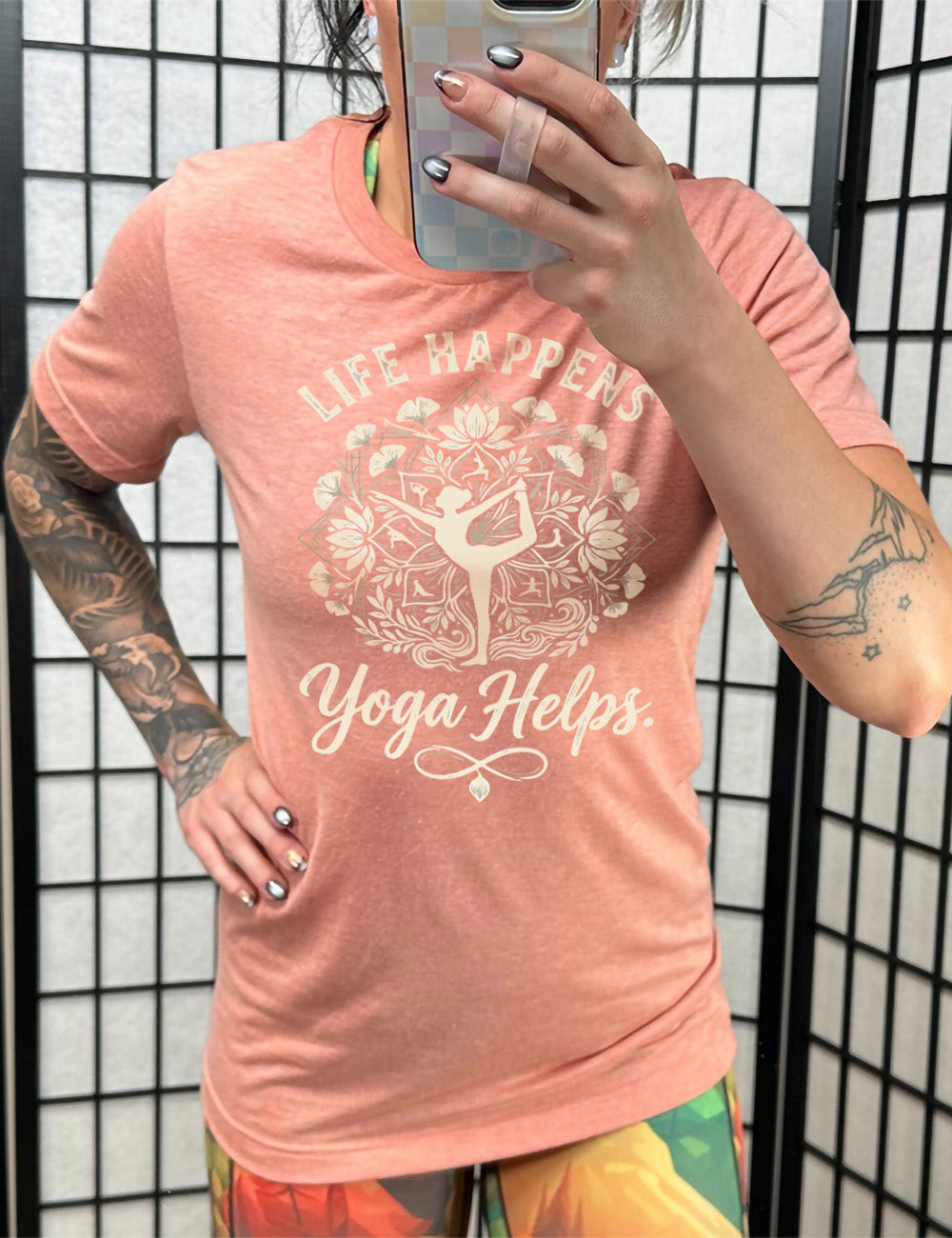 sunset unisex tee with the words "life happens yoga helps." on it, around the graphic of a woman's silhouette doing a yoga pose in the middle of a elaborate pattern of shapes, lotuses and other plants