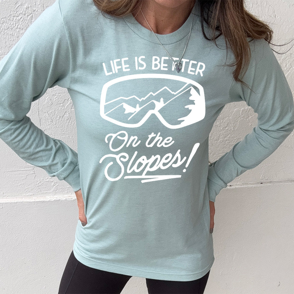 Heather dusty blue long sleeve with the words "Life is better on the slopes!" with the image of snow goggles and a reflection of the slopes in them