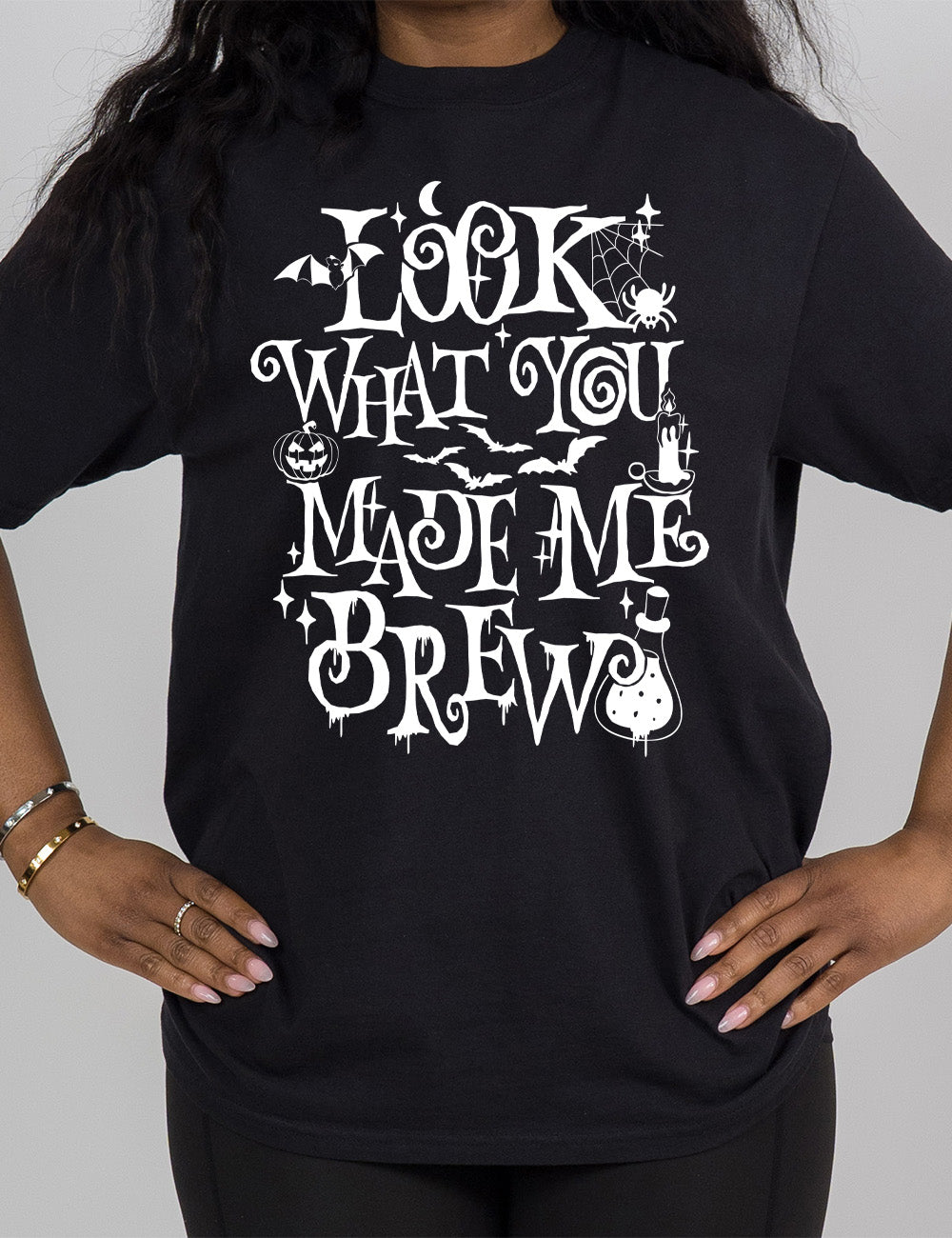 black modern tee with the curly, dripping words saying "look what you made me brew" (with the "o" being a spiral) and the graphics of a potion, a spider in its web, a candle, a bat some bats, stars, and a jack-o-lantern