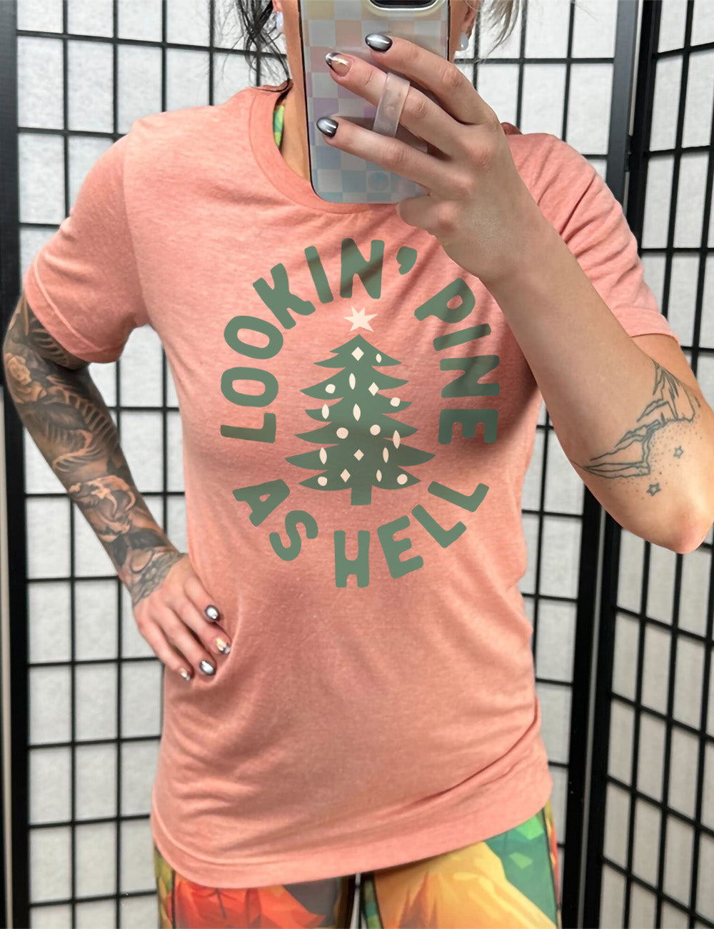 sunset unisex tee with the graphic of a Christmas tree, surrounded in oval shape by words saying "lookin' pine as hell" in capital letters