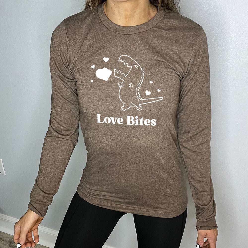 heather brown long sleeve shirt with the graphic of a dinosaur outline, eyes shut, mouth wide open, having bitten a chunk out of a heart, with a few other little hearts around it, and the text underneath saying "love bites"