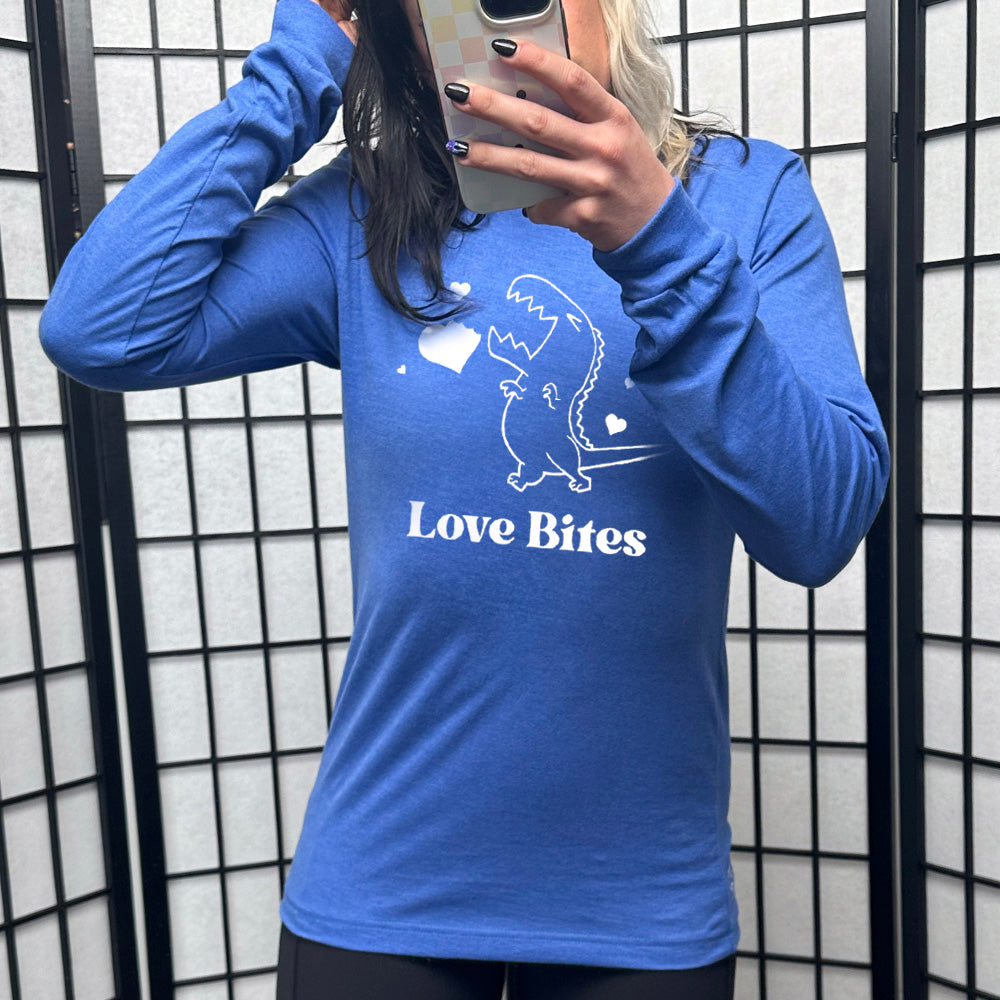 heather true royal long sleeve shirt with the graphic of a dinosaur outline, eyes shut, mouth wide open, having bitten a chunk out of a heart, with a few other little hearts around it, and the text underneath saying "love bites"