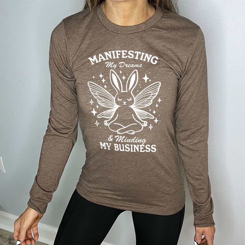 Heather brown long sleeve shirt with the words "Manifesting My Dreams & Minding My Business" and the graphic of a meditating rabbit with butterfly wings