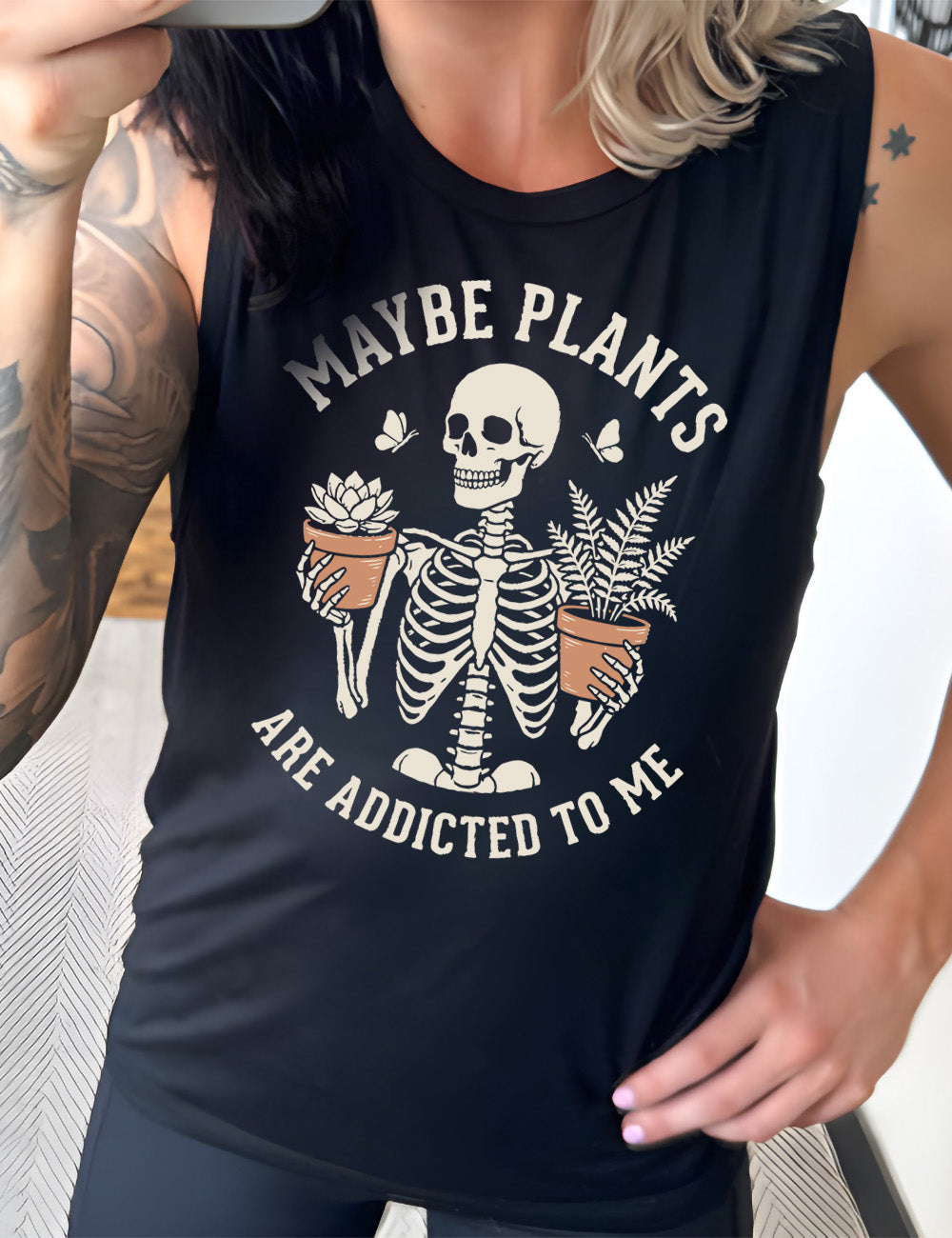 black muscle tank with the graphic of a skeleton, torso up, holding a houseplant in each hand, with the words around this saying "maybe plants are addicted to me"