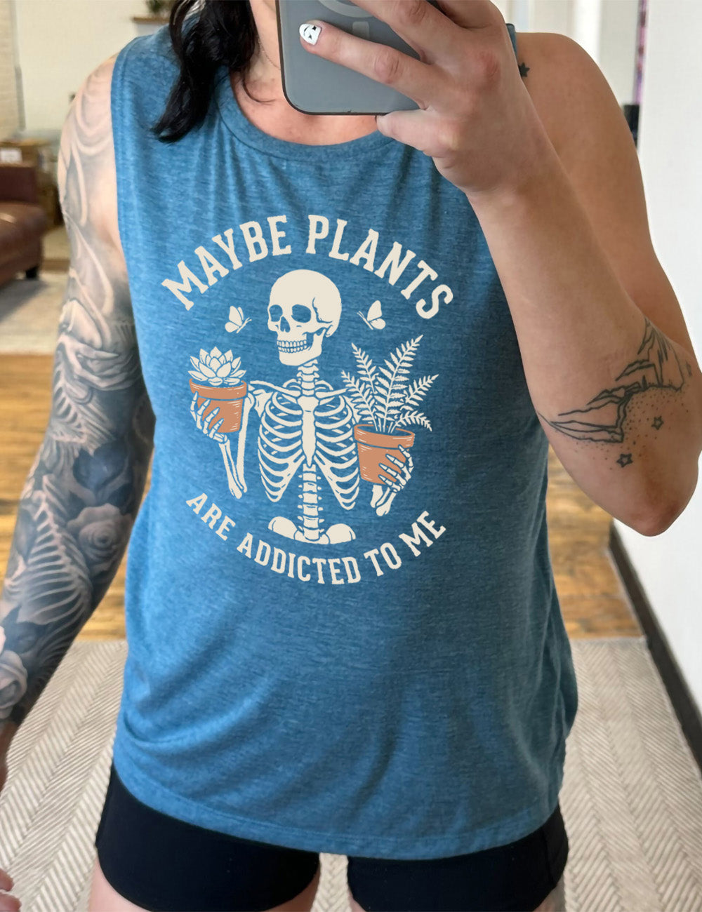 deep teal muscle tank with the graphic of a skeleton, torso up, holding a houseplant in each hand, with the words around this saying "maybe plants are addicted to me"